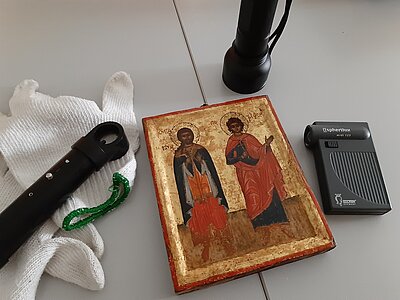 Eine Ikone mit zwei Heiligen, umgeben von Handschuhen, Lupe und zwei kleinen Taschenlampen