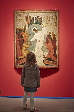 Eine Ikone mit der Hadesfahrt Christi hängt vor einem roten Hintergrund. Frontal vor der Ikone steht mit dem Rücken zum Betrachter ein junges Mädchen und betrachtet die Ikone interessiert.